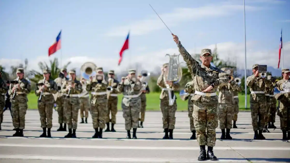 Un grupo de uniformados marcha en formación mientras un oficial al frente levanta un bastón de mando; al fondo se ven banderas chilenas y parte de una banda instrumental.