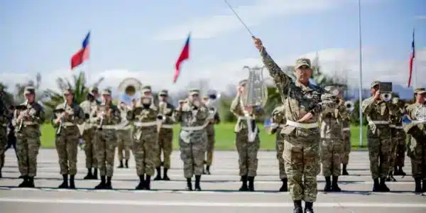 Un grupo de uniformados marcha en formación mientras un oficial al frente levanta un bastón de mando; al fondo se ven banderas chilenas y parte de una banda instrumental.