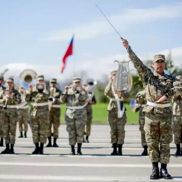 Un grupo de uniformados marcha en formación mientras un oficial al frente levanta un bastón de mando; al fondo se ven banderas chilenas y parte de una banda instrumental.