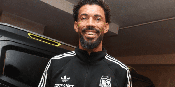 Hombre sonriente con chaqueta deportiva negra y blanca con el escudo de Colo Colo, de pie junto a un vehículo oscuro en un interior.