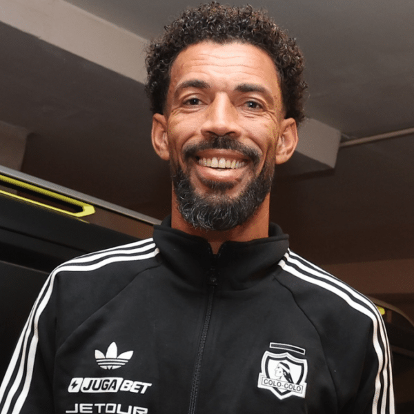 Hombre sonriente con chaqueta deportiva negra y blanca con el escudo de Colo Colo, de pie junto a un vehículo oscuro en un interior.