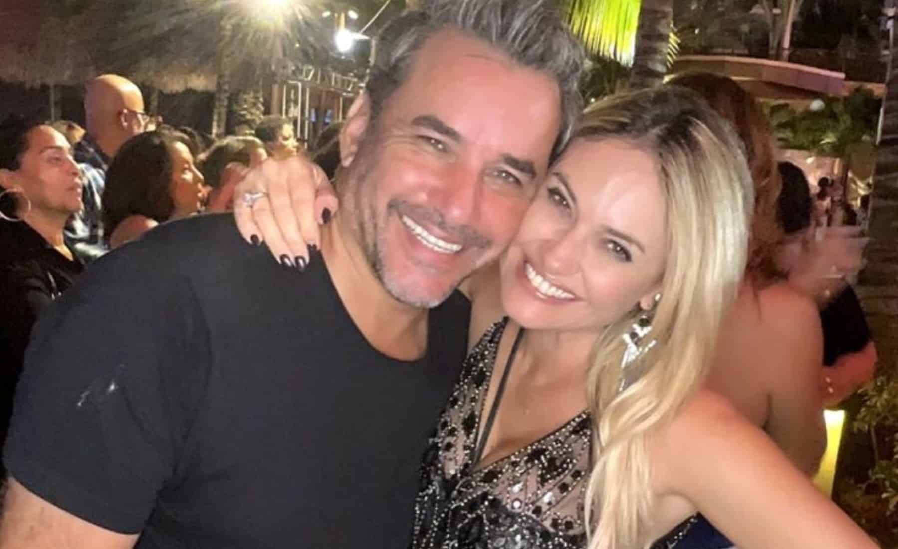 Un hombre de camiseta negra y una mujer rubia con vestido brillante posan abrazados y sonrientes en una celebración nocturna al aire libre, con luces y asistentes al fondo.