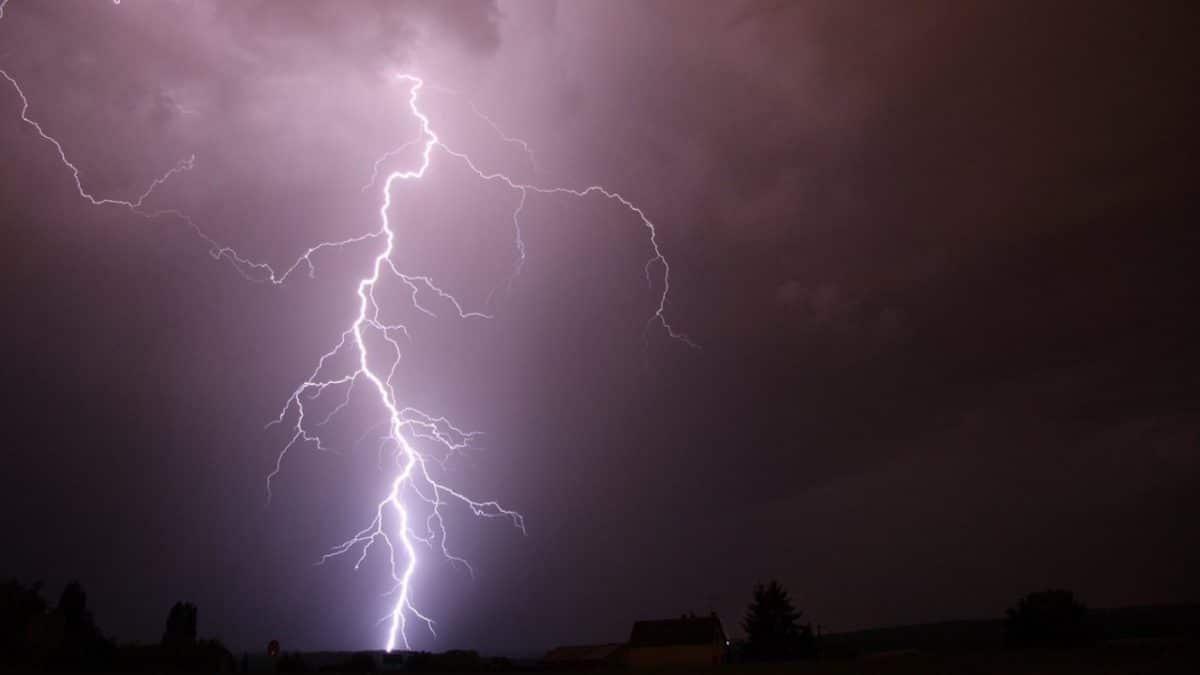 Un relámpago intenso cruza un cielo oscuro durante la noche, con siluetas de árboles y construcciones bajas en el horizonte.