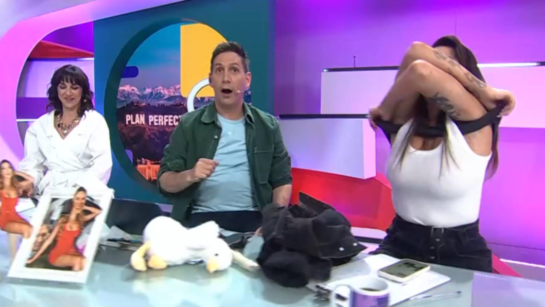 Panel de programa de televisión reacciona sorprendido mientras una mujer de pie se quita el sostén por encima de su polera blanca frente a las cámaras.