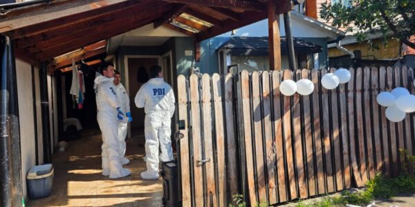Dos funcionarios de la PDI conversan en el acceso de una vivienda, junto a una reja de madera y globos blancos sujetos al cierre.