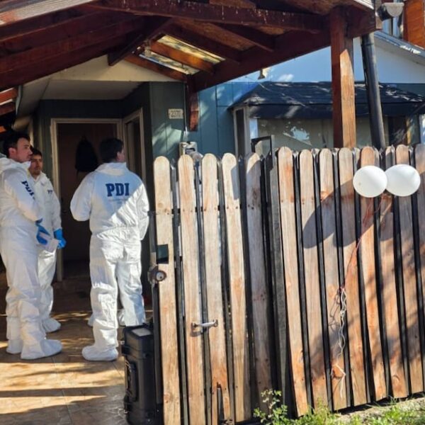 Dos funcionarios de la PDI conversan en el acceso de una vivienda, junto a una reja de madera y globos blancos sujetos al cierre.
