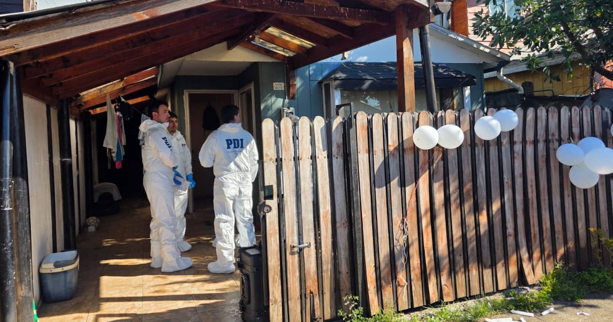 Dos funcionarios de la PDI conversan en el acceso de una vivienda, junto a una reja de madera y globos blancos sujetos al cierre.
