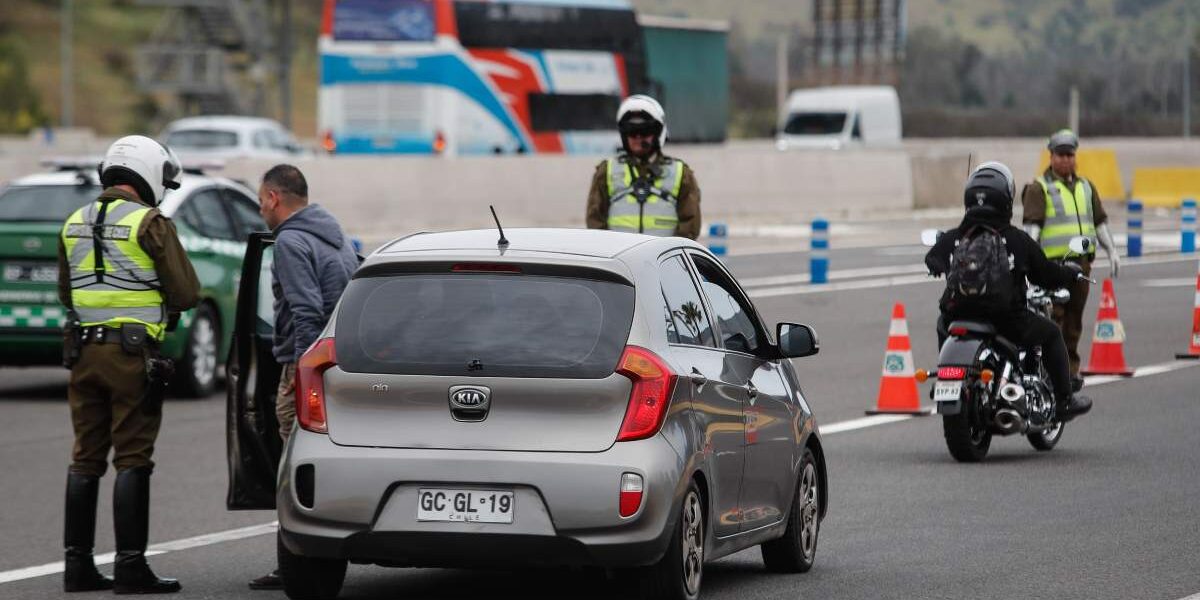 Carabineros fiscaliza a automovilistas en un control vial, con conos naranjos en la calzada, un auto detenido al centro y un motociclista avanzando por la vía.