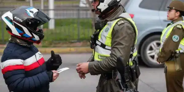 Carabinero con chaleco reflectante conversa con una persona que lleva casco de moto y sostiene un documento, mientras otro uniformado permanece al fondo junto a un vehículo.