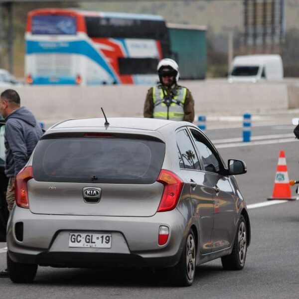Carabineros fiscaliza a automovilistas en un control vial, con conos naranjos en la calzada, un auto detenido al centro y un motociclista avanzando por la vía.