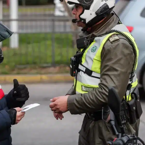 Carabinero con chaleco reflectante conversa con una persona que lleva casco de moto y sostiene un documento, mientras otro uniformado permanece al fondo junto a un vehículo.