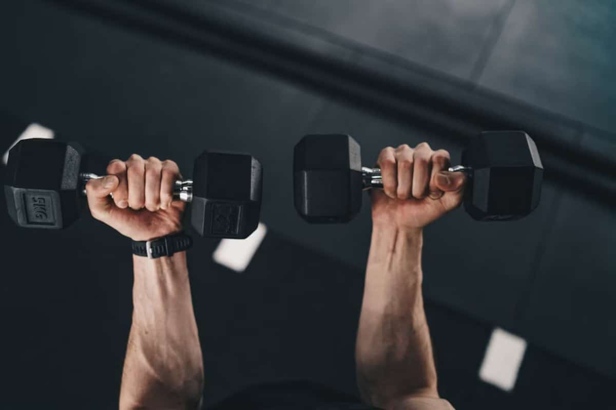 Primer plano de dos manos sosteniendo mancuernas negras levantadas sobre el fondo oscuro de un gimnasio.