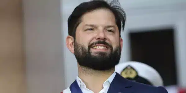 Hombre con traje azul y banda tricolor sonríe durante una ceremonia, con fondo desenfocado y una persona uniformada detrás.
