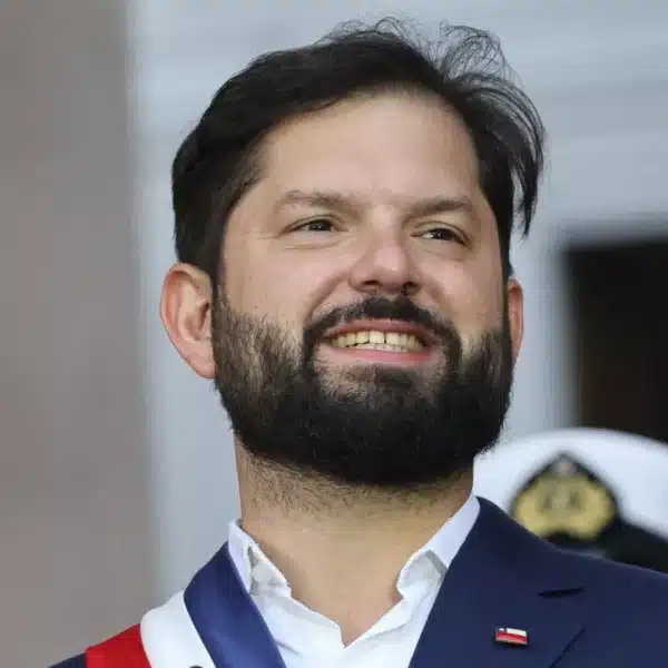 Hombre con traje azul y banda tricolor sonríe durante una ceremonia, con fondo desenfocado y una persona uniformada detrás.