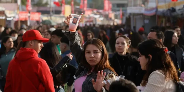 Personas reunidas en una fonda al aire libre, con banderas chilenas al fondo. En primer plano, una mujer levanta un vaso mientras conversa con otras personas entre la multitud.