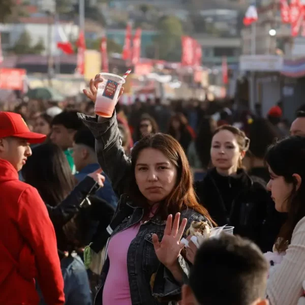 Personas reunidas en una fonda al aire libre, con banderas chilenas al fondo. En primer plano, una mujer levanta un vaso mientras conversa con otras personas entre la multitud.