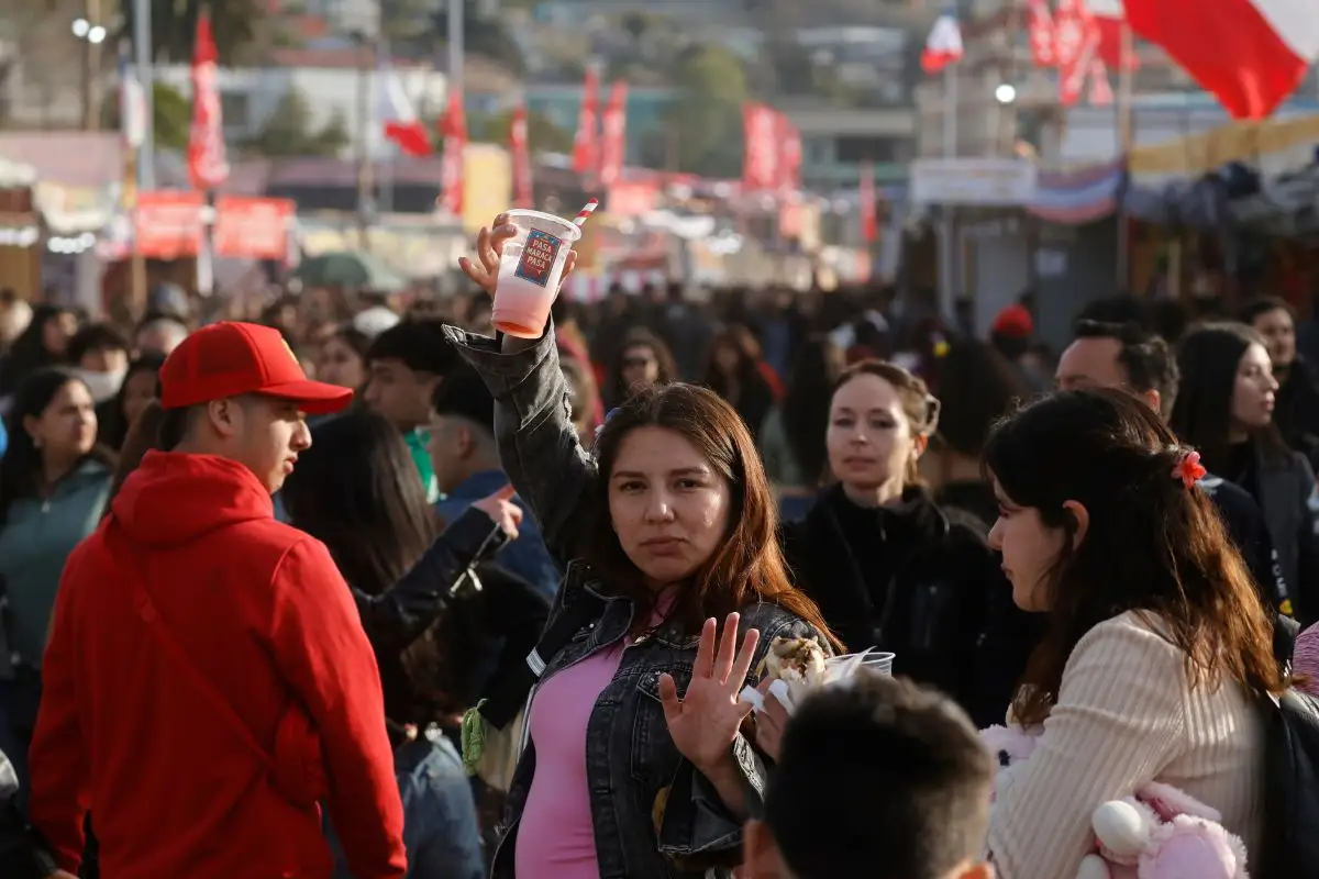 Personas reunidas en una fonda al aire libre, con banderas chilenas al fondo. En primer plano, una mujer levanta un vaso mientras conversa con otras personas entre la multitud.