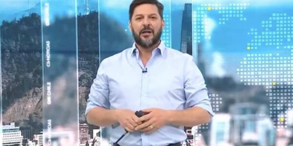 Un hombre de pie en un estudio de televisión, con camisa celeste y micrófono de solapa, frente a un fondo gráfico azul con imágenes urbanas y paneles luminosos.