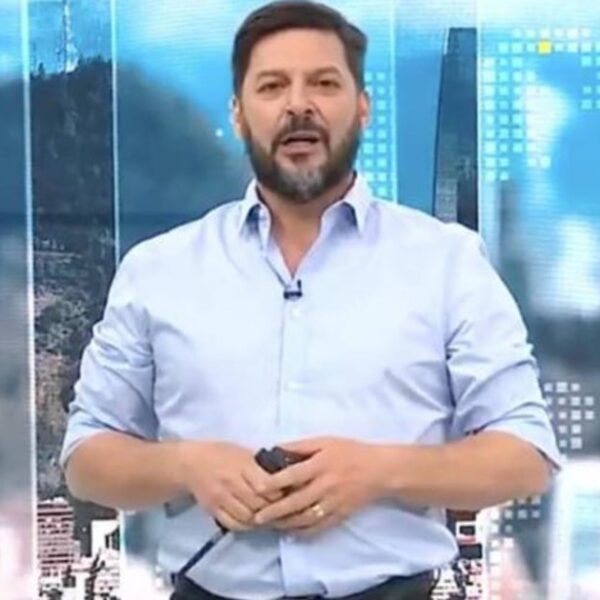 Un hombre de pie en un estudio de televisión, con camisa celeste y micrófono de solapa, frente a un fondo gráfico azul con imágenes urbanas y paneles luminosos.