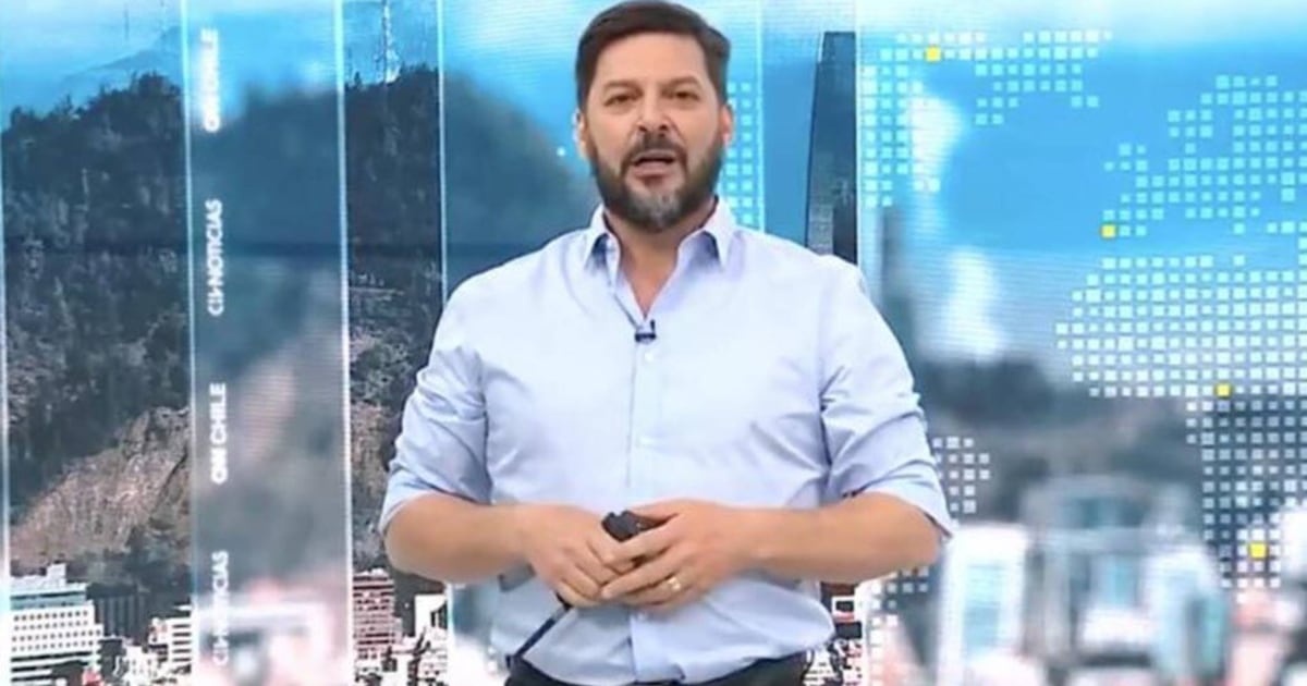 Un hombre de pie en un estudio de televisión, con camisa celeste y micrófono de solapa, frente a un fondo gráfico azul con imágenes urbanas y paneles luminosos.