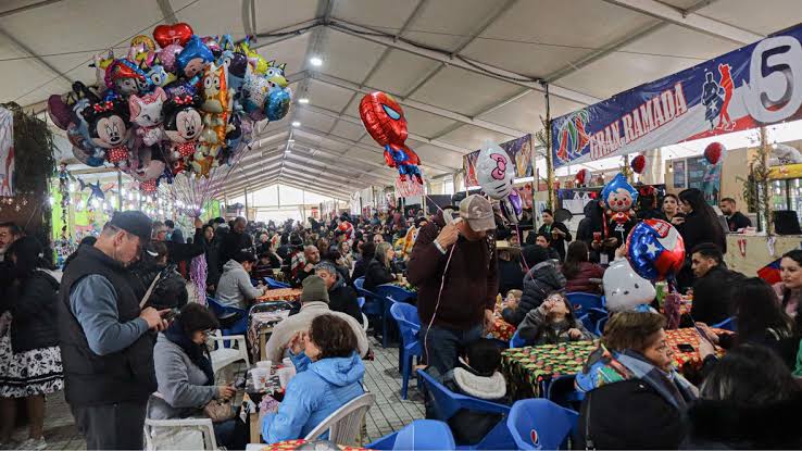 Interior de una fonda bajo carpa, con muchas personas sentadas en mesas azules comiendo y conversando, mientras al fondo se ven puestos decorados y globos de colores.