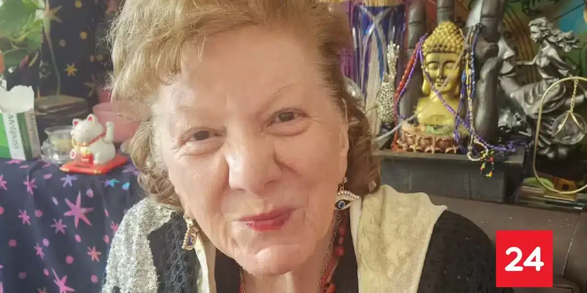 Mujer de cabello claro sonríe a la cámara en un interior lleno de objetos decorativos, con una figura de Buda y adornos al fondo.