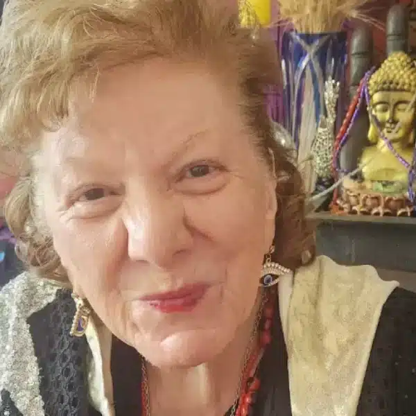 Mujer de cabello claro sonríe a la cámara en un interior lleno de objetos decorativos, con una figura de Buda y adornos al fondo.