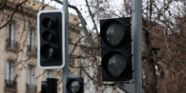 Semáforos vehiculares y peatonales apagados en postes de una calle, con edificios y árboles sin hojas al fondo.