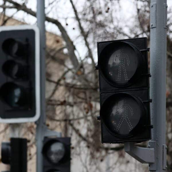Semáforos vehiculares y peatonales apagados en postes de una calle, con edificios y árboles sin hojas al fondo.