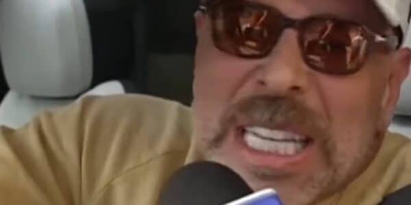 Primer plano de un hombre con gorra clara y lentes oscuros, hablando con expresión intensa frente a un micrófono.