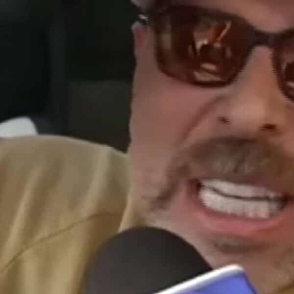 Primer plano de un hombre con gorra clara y lentes oscuros, hablando con expresión intensa frente a un micrófono.