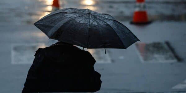 Una persona de espaldas sostiene un paraguas negro en una calle mojada, con lluvia y conos de tránsito desenfocados al fondo.