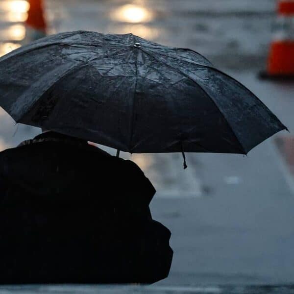 Una persona de espaldas sostiene un paraguas negro en una calle mojada, con lluvia y conos de tránsito desenfocados al fondo.