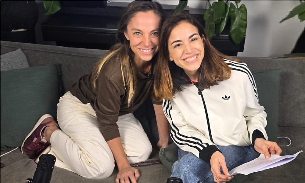 Dos mujeres sonrientes sentadas en el suelo junto a un sofá gris; una viste buzo café y pantalón blanco, la otra chaqueta deportiva blanca y jeans, sosteniendo papeles.