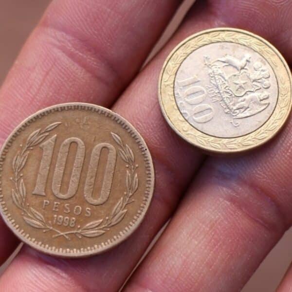 Varias monedas de $100 chilenas sobre una mesa junto a un vuelto de compra, piezas que pueden alcanzar altos valores entre coleccionistas numismáticos.