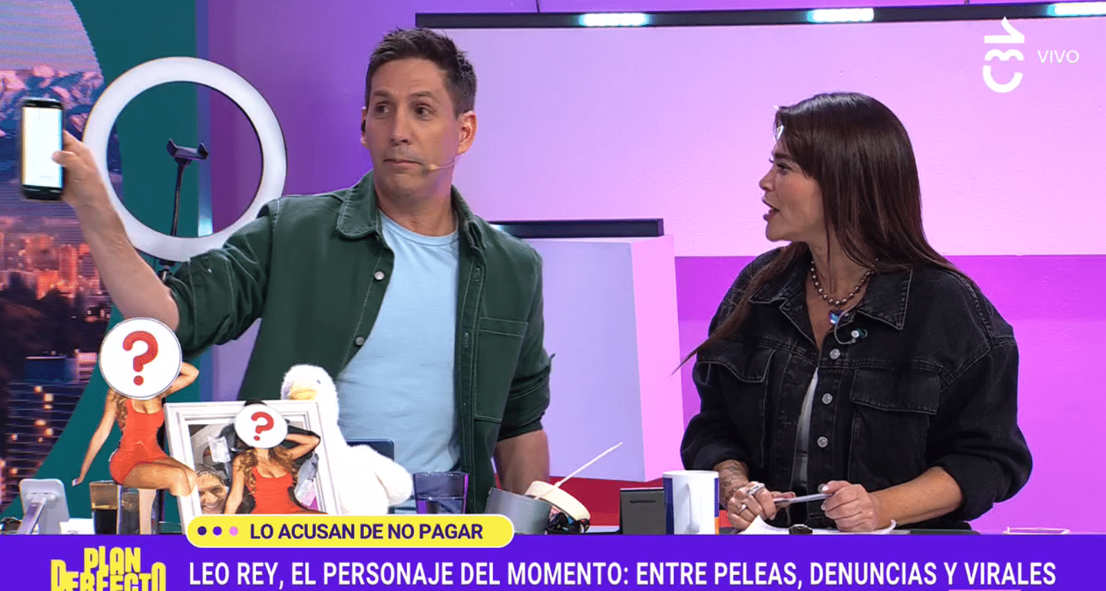 Dos panelistas de un programa de televisión en vivo: un hombre con chaqueta verde muestra su teléfono celular al aire mientras una mujer con chaqueta negra lo mira, con gráficas alusivas a Leo Rey en pantalla.