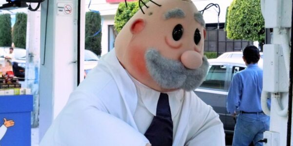 Una persona disfrazada de Dr. Simi, con bata blanca y corbata oscura, aparece en la entrada de un local, con autos y personas al fondo.