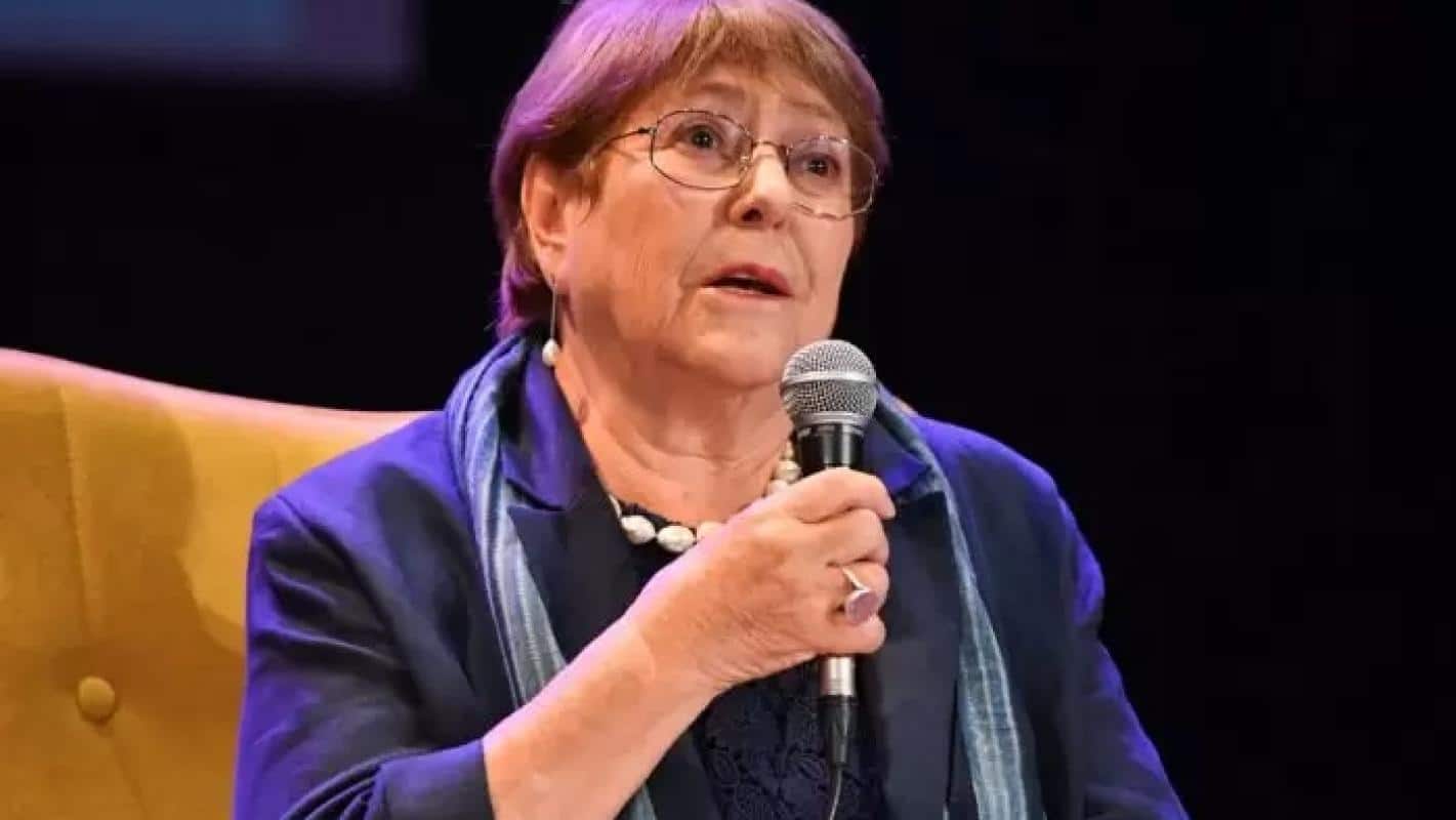 Mujer mayor con lentes y chaqueta azul sostiene un micrófono mientras habla sentada en un sillón amarillo sobre un fondo oscuro.