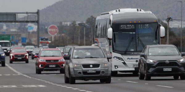Varios autos y un bus circulan por una autopista de varias pistas, con tráfico en movimiento y un cerro de fondo bajo cielo nublado.
