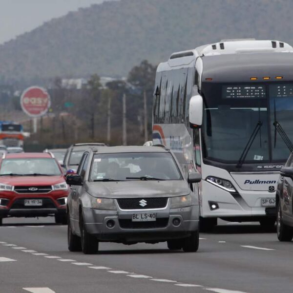 Varios autos y un bus circulan por una autopista de varias pistas, con tráfico en movimiento y un cerro de fondo bajo cielo nublado.