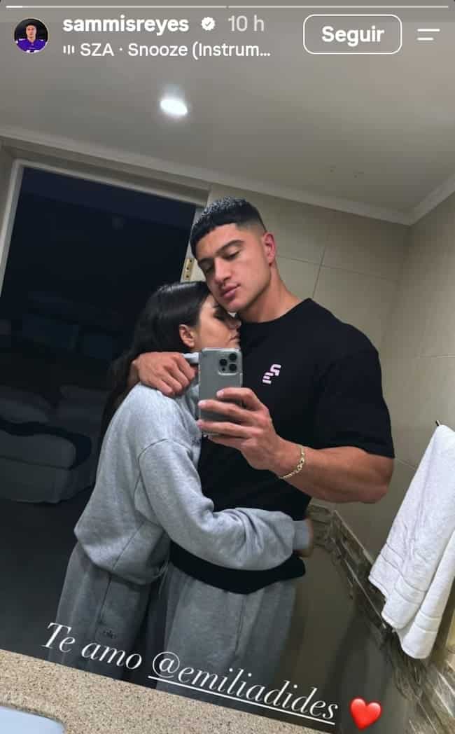 Una pareja se toma una selfie frente al espejo en un baño, abrazada con gesto cariñoso. Él viste polera negra y ella buzo gris; abajo se ve un mensaje de amor con un emoji de corazón.