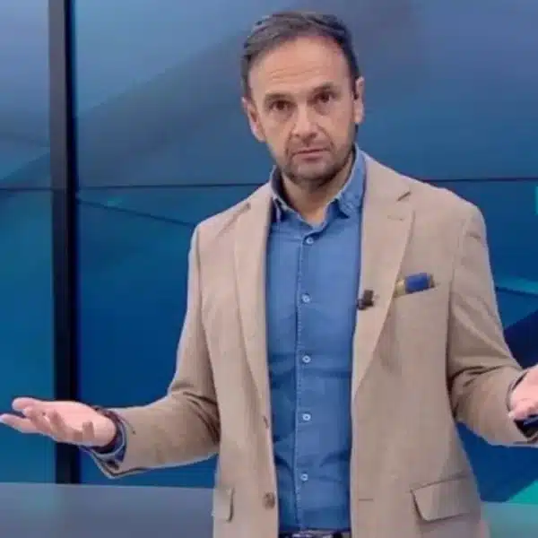 Hombre con blazer beige y camisa azul habla con las palmas abiertas en un estudio azul; al fondo se ven pantallas con un símbolo de hashtag y gráficos.
