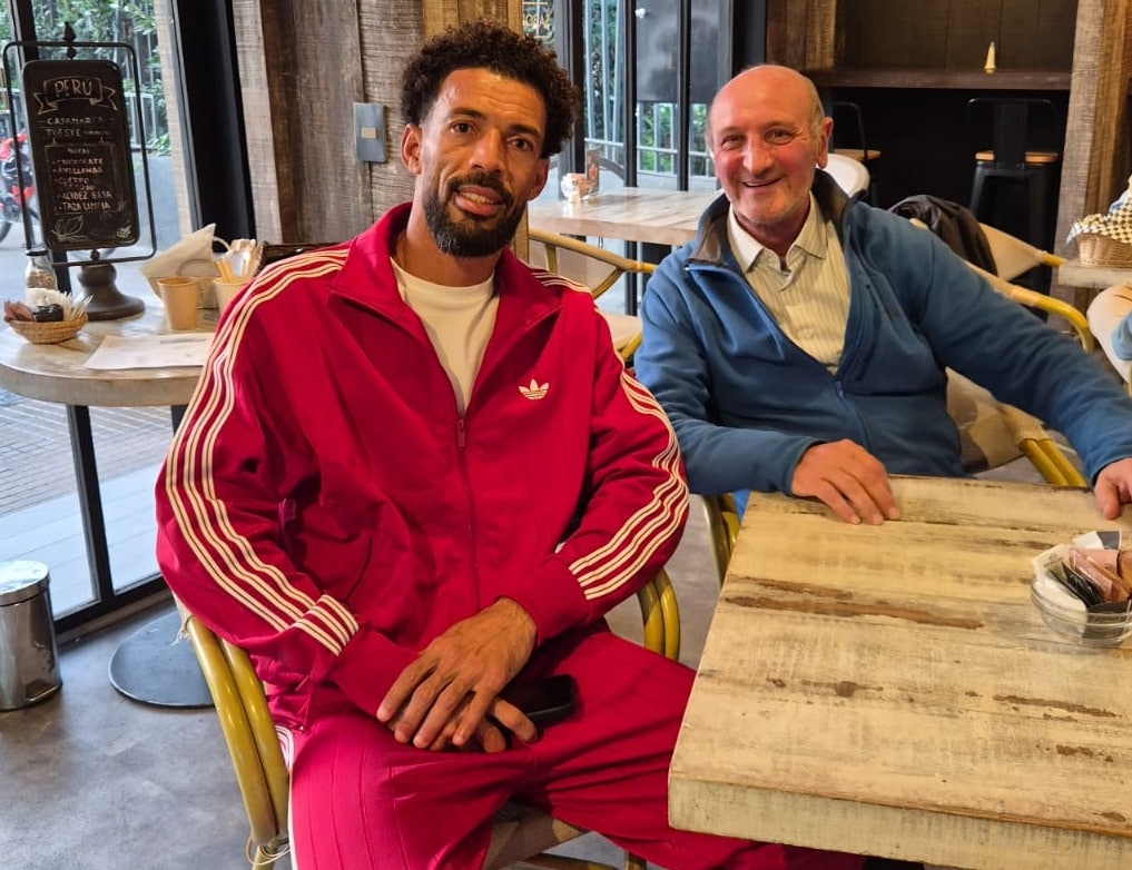 Dos hombres están sentados en una cafetería, uno con buzo deportivo rojo y otro con chaqueta azul, junto a una mesa de madera en un ambiente interior iluminado.