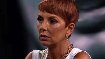 Primer plano de una mujer de pelo corto rojizo, con top blanco sin mangas y collares dorados, mirando hacia un costado con expresión seria ante un fondo oscuro difuminado.