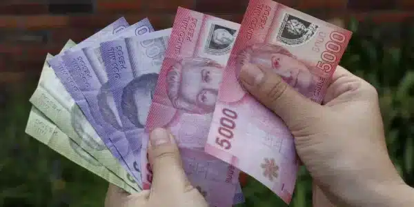 Dos manos sostienen varios billetes chilenos de distintos colores y denominaciones, con billetes de 5000 pesos visibles al frente.
