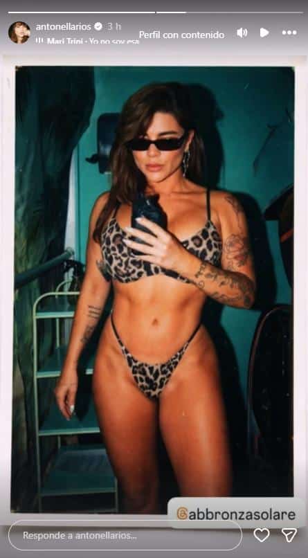 Una mujer con tatuajes y lentes de sol posa de pie con un bikini estampado en un espacio interior.