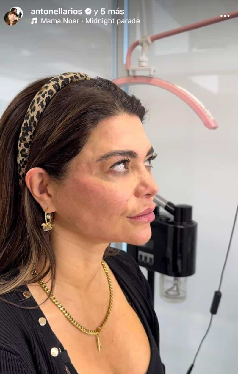 Una mujer de perfil mira hacia arriba. Lleva un cintillo estampado, aros dorados y una blusa negra; detrás se ven equipos y una lámpara.