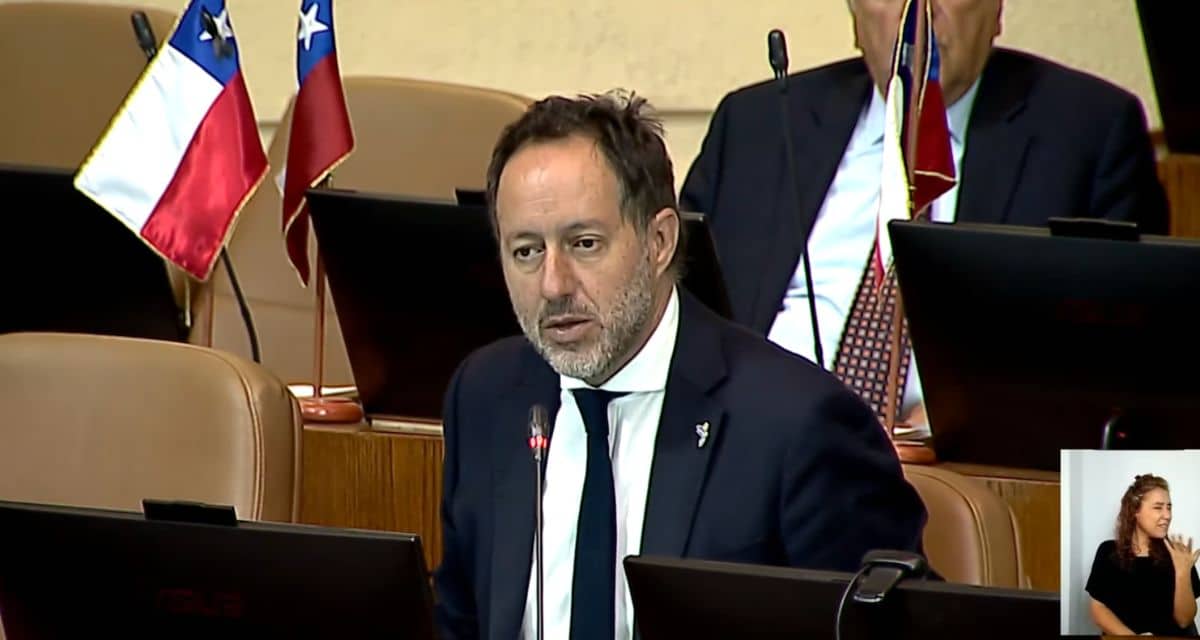 Un hombre habla ante un micrófono en una sala con escaños y banderas chilenas; en una ventana al costado aparece una intérprete de lengua de señas.