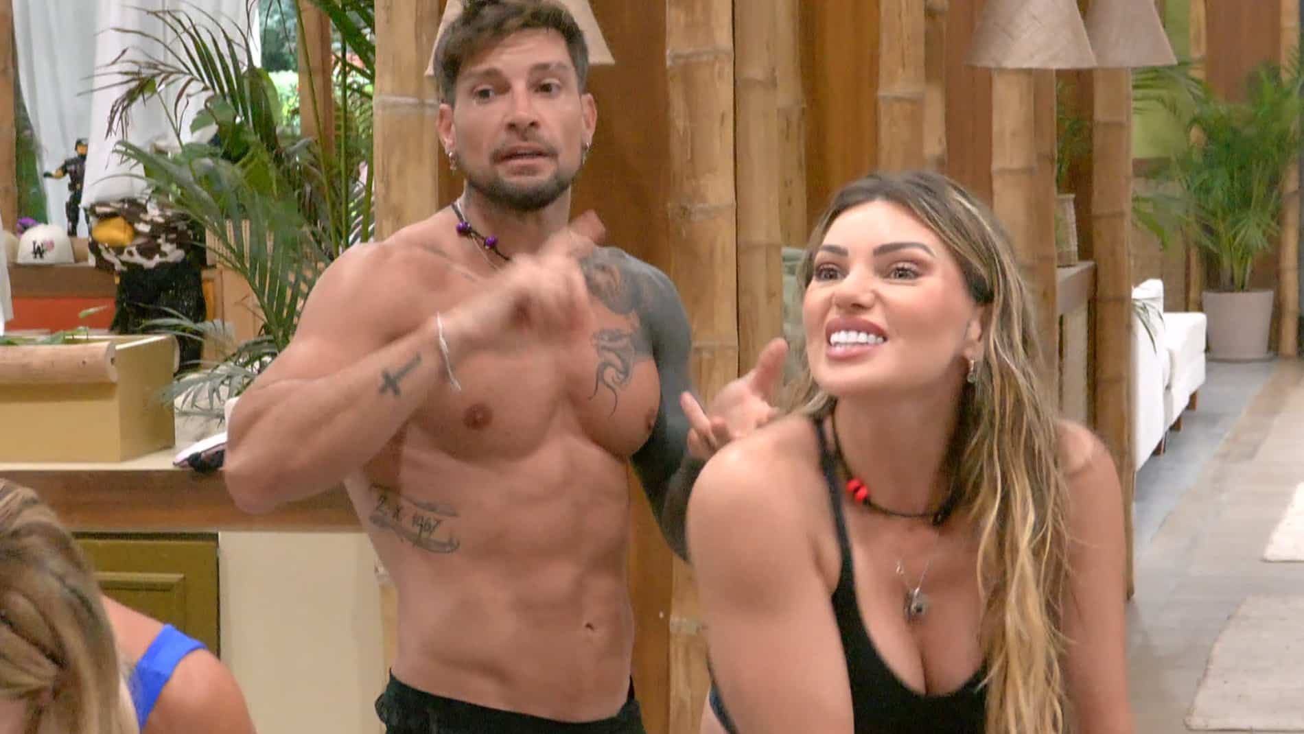 Un hombre sin polera, con tatuajes, gesticula junto a una mujer que sonríe y se inclina hacia adelante, en una habitación de madera.