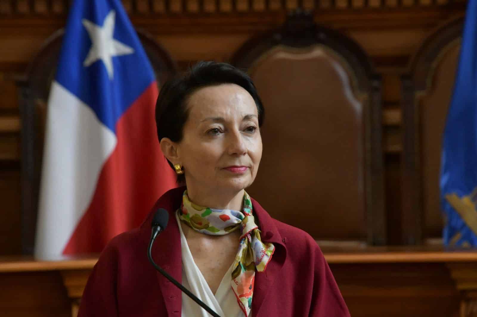 Una mujer con chaqueta burdeos y pañuelo estampado está frente a un micrófono, con una bandera chilena y mobiliario de madera al fondo.